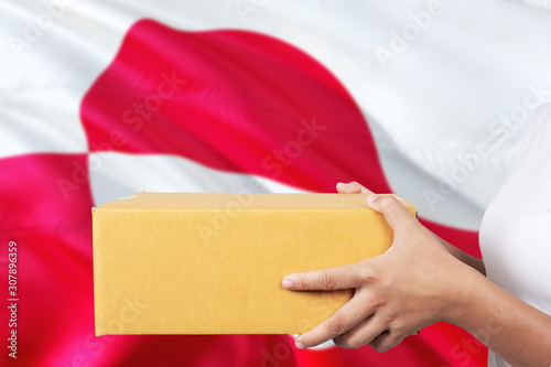 Greenland delivery service. International shipment theme. Woman courier hand holding brown box isolated on national flag background.