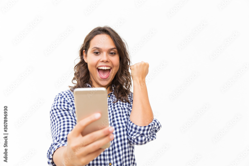 Excited Latin woman with cellphone getting good news and making winner gesture. Young woman in casual checked shirt standing isolated over white background. Winning prize concept Excited Latin woman with cellphone getting good news and making winner gesture. Young woman in casual checked shirt standing isolated over white background. Winning prize concept