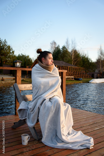 A young girl in a warm plaid rests by the lake in autumn. The concept of enjoying nature, relaxation, travel © Uladzimir
