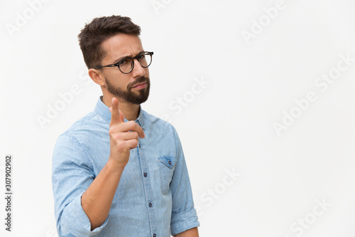Frowning concerned guy shaking finger at camera. Handsome young man in casual shirt and glasses standing isolated over white background. Warning gesture concept