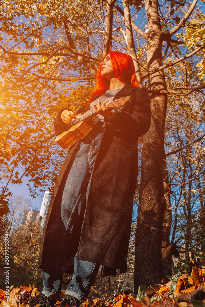 Red-haired girl with long hair plays on the ukulele in the park. School, music education concept, student learns to play the string instrument. Hands of a musician, classical, melody, creativity. Red-haired girl with long hair plays on the ukulele in the park. School, music education concept, student learns to play the string instrument. Hands of a musician, classical, melody, creativity.