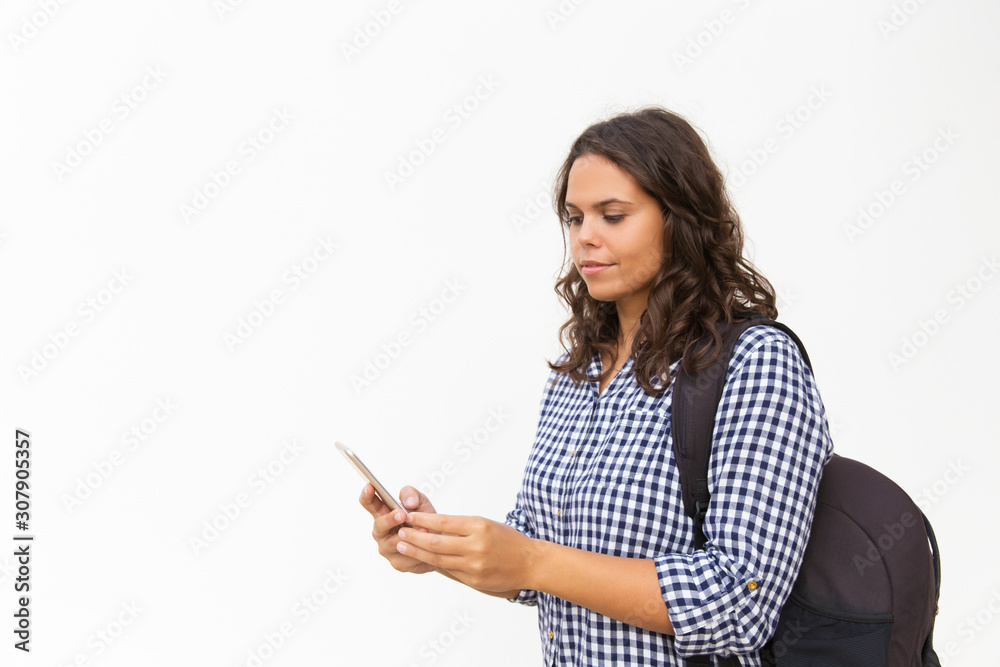 Focused female tourist with backpack and cellphone using location app. Young woman in casual checked shirt standing isolated over white background. Tourist with smartphone concept Focused female tourist with backpack and cellphone using location app. Young woman in casual checked shirt standing isolated over white background. Tourist with smartphone concept