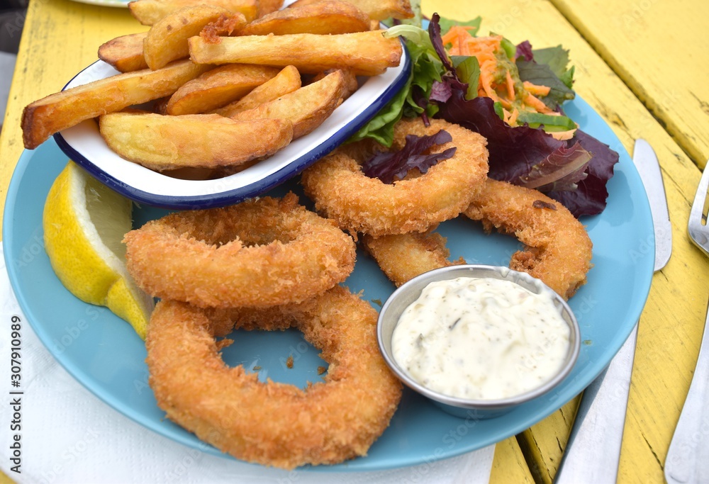 Fried calamari with salad and potato chips on blue ceramic plate and yellow table background. Fried calamari with salad and potato chips on blue ceramic plate and yellow table background.