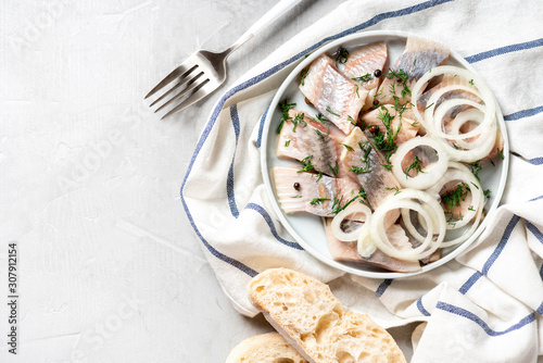 Pieces of pickled herring with onions, dill and pepper in a ceramic plate on a light gray background, top view, copy space.