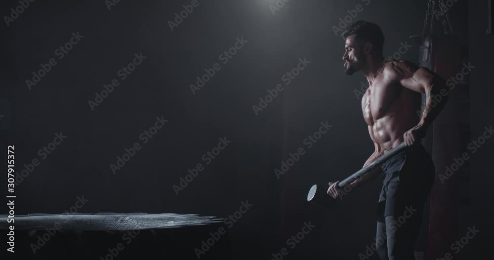 Charismatic guy with a muscle body strikes over the tire using a hummer with full of power he have a angry and concentrated face Charismatic guy with a muscle body strikes over the tire using a hummer with full of power he have a angry and concentrated face