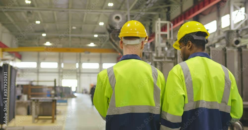 Two factory workers walking with discussion holding tablet pc. Industrial heavy industry enterprise background. Foremen in helmets and uniform walking through factory facility