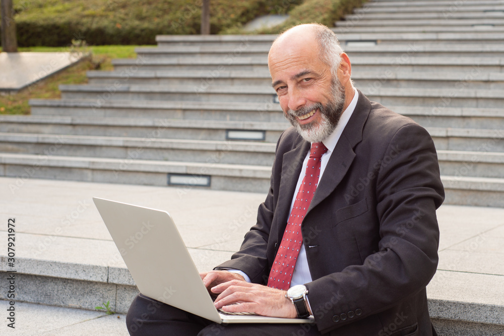 Businessman using laptop and smiling at camera. Cheerful mature businessman in formal wear sitting on steps and working with laptop computer. Business and technology concept Businessman using laptop and smiling at camera. Cheerful mature businessman in formal wear sitting on steps and working with laptop computer. Business and technology concept