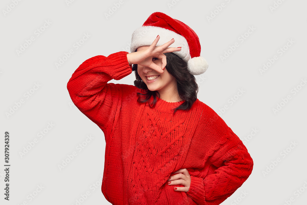 Pretty brunette girl in a red sweater and Santa hat showing OK sign isolated over grey background. Pretty brunette girl in a red sweater and Santa hat showing OK sign isolated over grey background.
