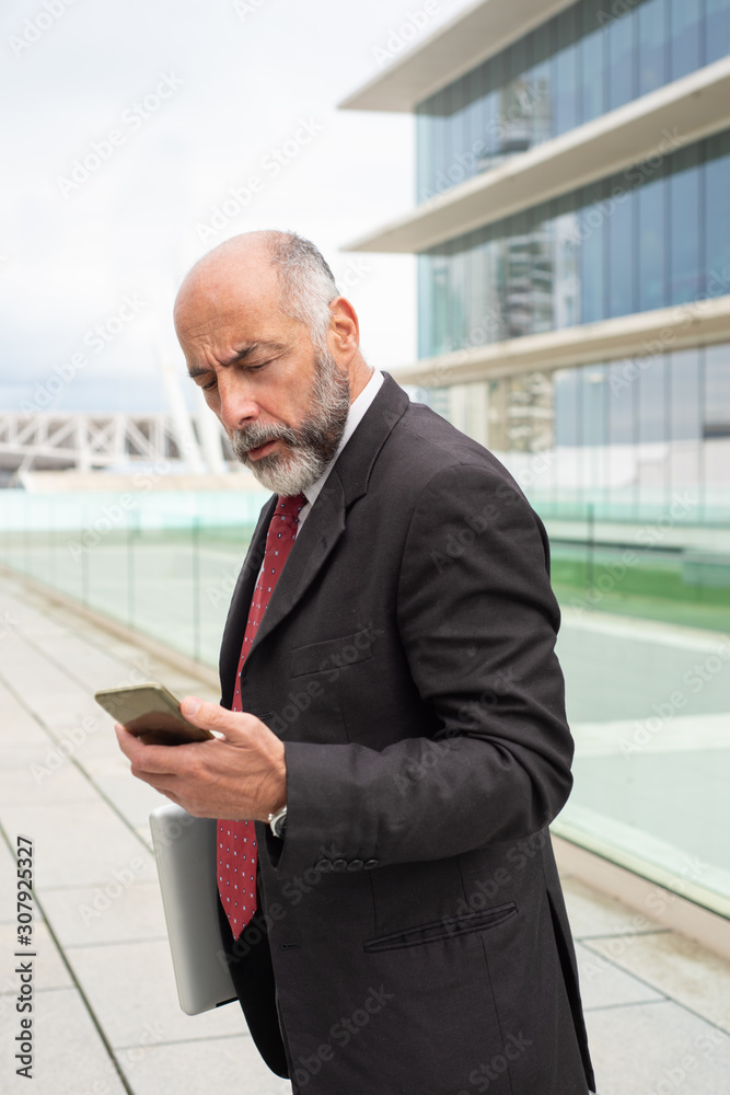 Focused mature business leader with laptop reading message on smartphone. Elderly man in formal suit and tie standing outside in city. Communication concept Focused mature business leader with laptop reading message on smartphone. Elderly man in formal suit and tie standing outside in city. Communication concept