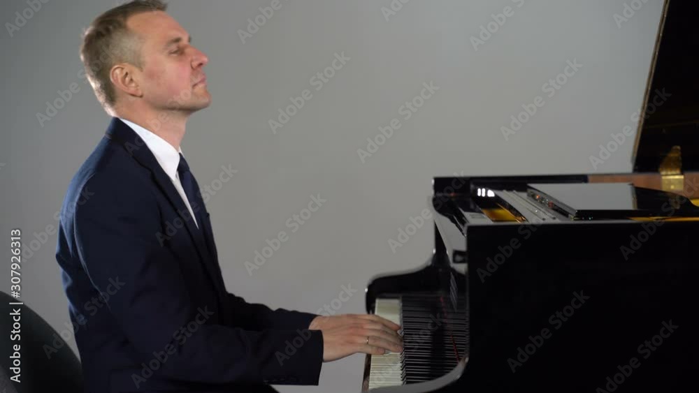 A man in a black suit plays the piano against a grey background
