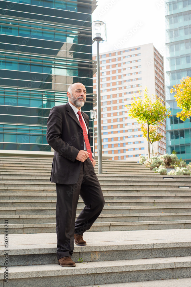 Mature businessman walking down stairs. Low angle view of confident bearded businessman in formal wear walking on stairs and looking aside on urban city street. Business concept Mature businessman walking down stairs. Low angle view of confident bearded businessman in formal wear walking on stairs and looking aside on urban city street. Business concept