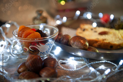 Antique, transparent cup, dried fruits. Focused damasco. Brazilian Christmas table. Portuguese chestnut, panettone, defocused. Selective focus. White Christmas Lights. Seen up close. Horizontal