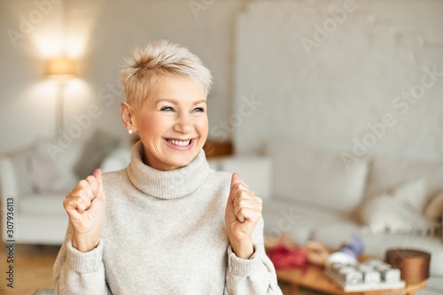 Photography Indoor shot of fashionable overjoyed mature female in turtleneck sweater enjoying positive news, having ecstatic facial expression, laughing and clenching fists