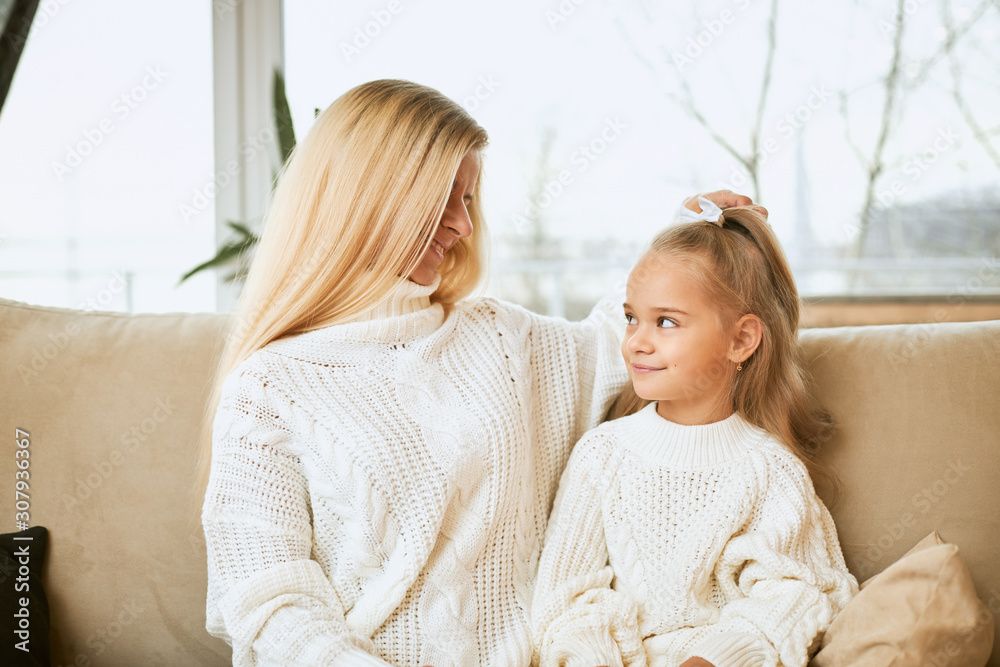 Cute adorable female child sitting on sofa with her young mother. Blonde woman in white cozy sweater stroking hair of beautiful daughter, cuddling on couch in living room. People and generations Cute adorable female child sitting on sofa with her young mother. Blonde woman in white cozy sweater stroking hair of beautiful daughter, cuddling on couch in living room. People and generations