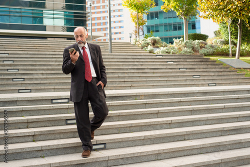 Businessman walking on steps and using smartphone. Full length view of bearded mature businessman using mobile phone on street. Technology concept