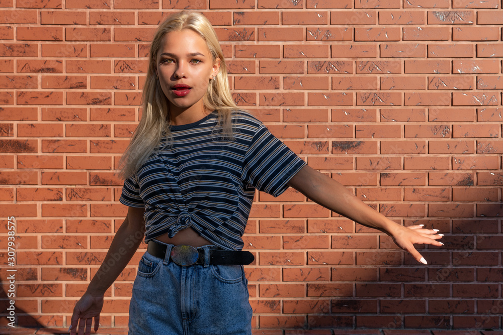 Attractive young blonde girl with red lips dressed in grey striped t-shirt withopen hands looking at the camera isolated over red brick wall