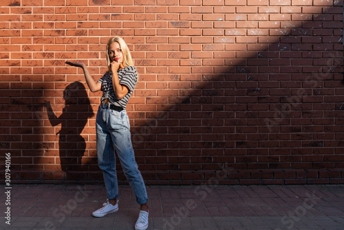 Thoughtful blonde girl in grey t-shirt and blue jeans shows, holds your prodact on the open palm isolated over red brick wall