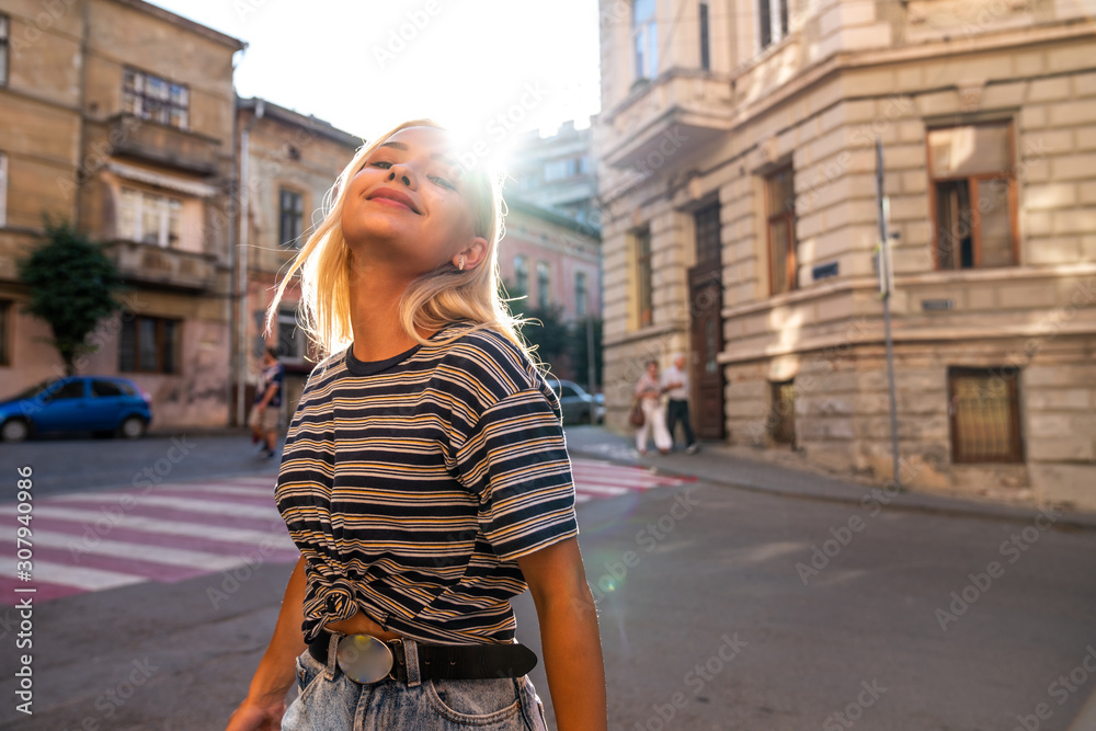 Waist up portrait of the young teen blonde girl with loose hair dressed in grey striped t-shirt walks among the old city center, sunset on the background