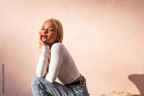 Beautiful blonde teen girl in white top crouched, leaned on her hands and looking at the camera, pink wall on the background
