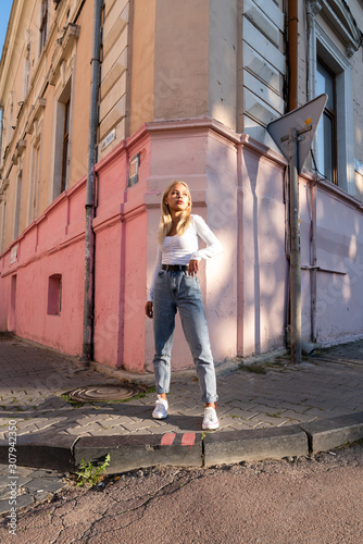 Street style portrait of the beautiful blonde teen girl in white top and blue jeans stands near the corner of the pink old house