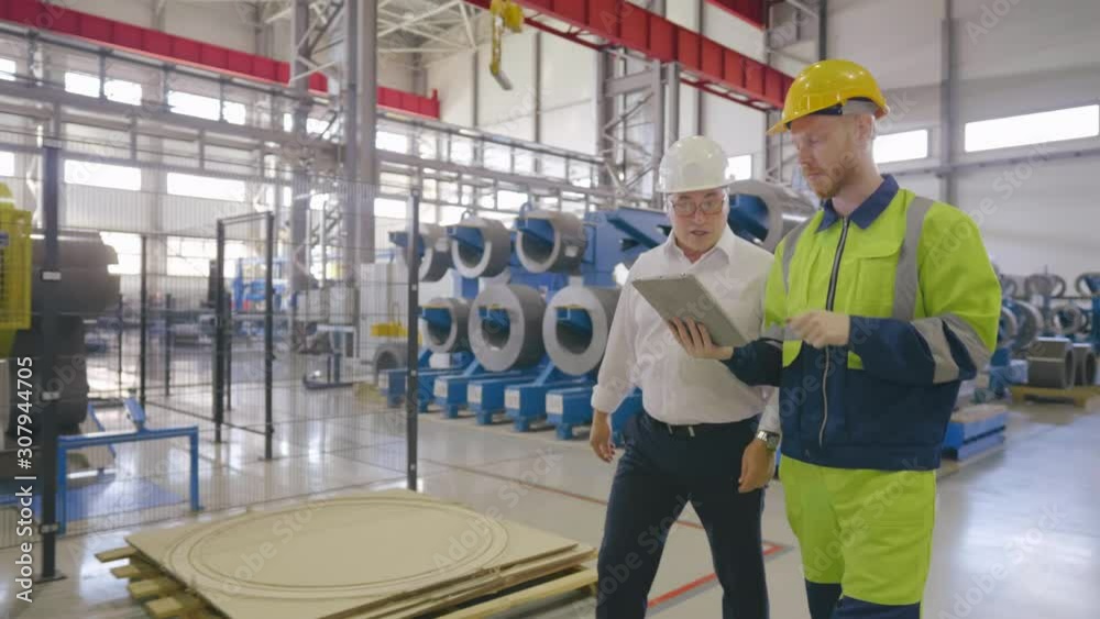 Mature engineer in white helmet and worker in uniform walk through facility of modern plant and discuss data project in tablet pc. Planning inside a big manufactory. Successful business