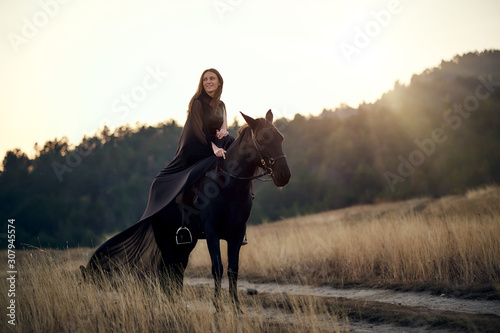 cheerful smiling young beautiful girl in a light flying black dress riding a black horse rides on the field against the background of the forest and sunset © MikeFrame