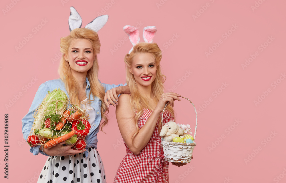 Young pin up women tweens in retro clothes with bunny ears holding a basket  full of Easter eggs and string bag with vegetables on pink background. Eco- friendly packaging and zero waste Stock