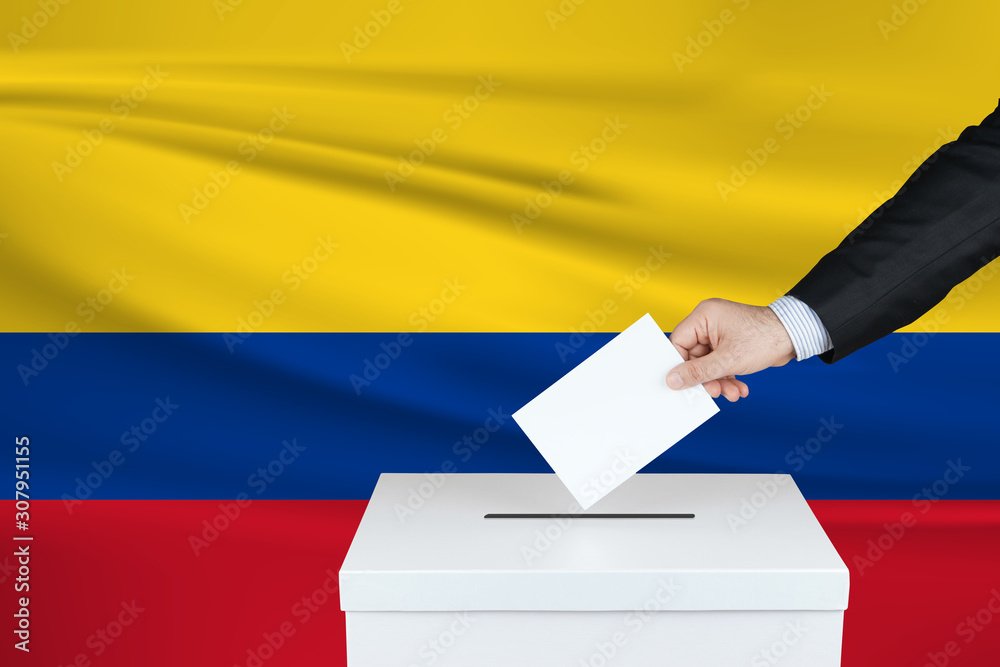 Election in Columbia. The hand of man putting his vote in the ballot box. Waved Columbia flag on background.