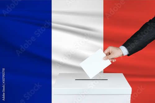 Election in France. The hand of man putting his vote in the ballot box. Waved France flag on background.