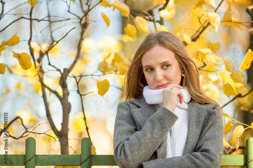 Beautiful girl with light brown hair near the fence and bushes with yellow foliage in the autumn park. Attractive young woman in a gray coat looks away. Fall season. Golden leaves. Amazing autumn mood