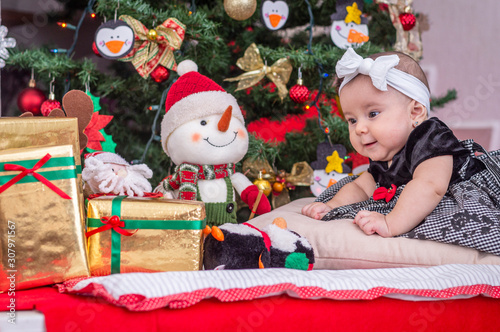 A white baby in Christmas tree decoration with black dress and white bow leaning on her hand looks at happy gifts