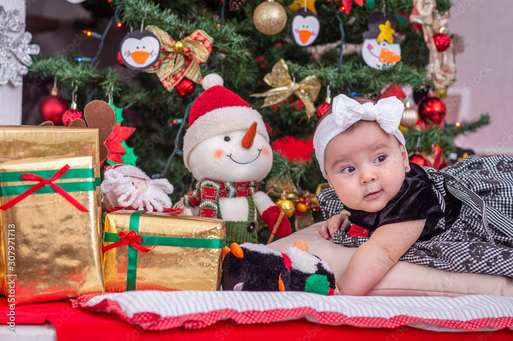 A white baby in Christmas tree decoration with black dress and white bow lying down looks very attentive A white baby in Christmas tree decoration with black dress and white bow lying down looks very attentive