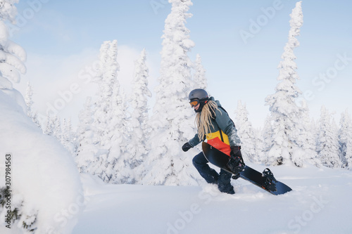 Female snowboarder wearing long dreadlocks and ratsa hoody walking between snow cowered pine trees in sunny winter day