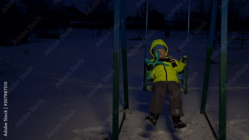 cute child in bright yellow jacket rides on swing on winter evening. Snowy winter in village. boy has fun alone in playground. Depressive evening lighting cute child in bright yellow jacket rides on swing on winter evening. Snowy winter in village. boy has fun alone in playground. Depressive evening lighting