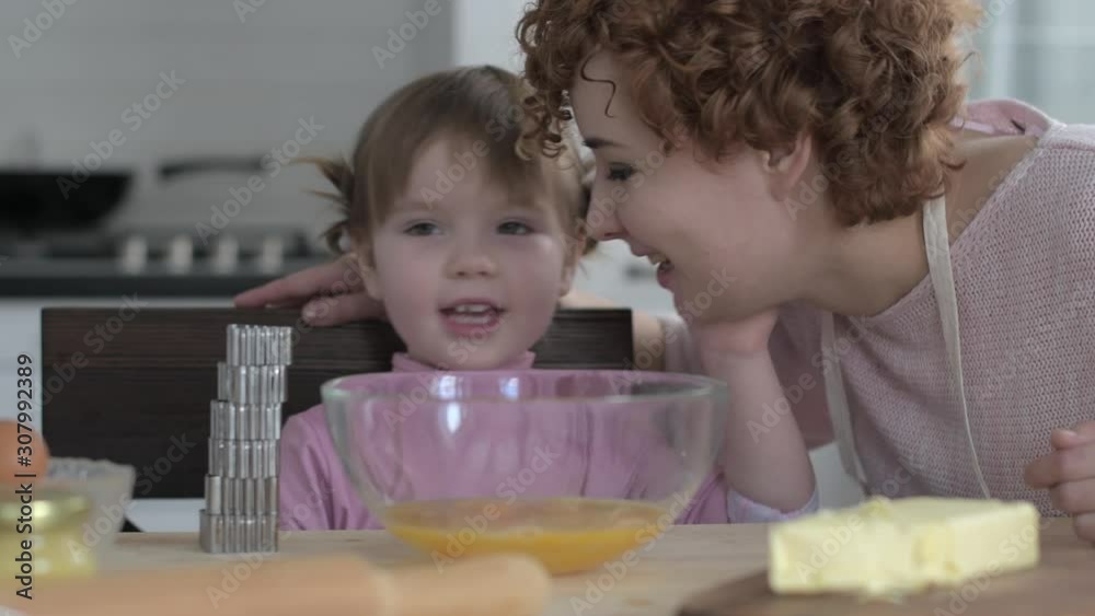 Daughter whispers to mom in the ear secrets while sitting in the kitchen for cooking. Daughter whispers to mom in the ear secrets while sitting in the kitchen for cooking.