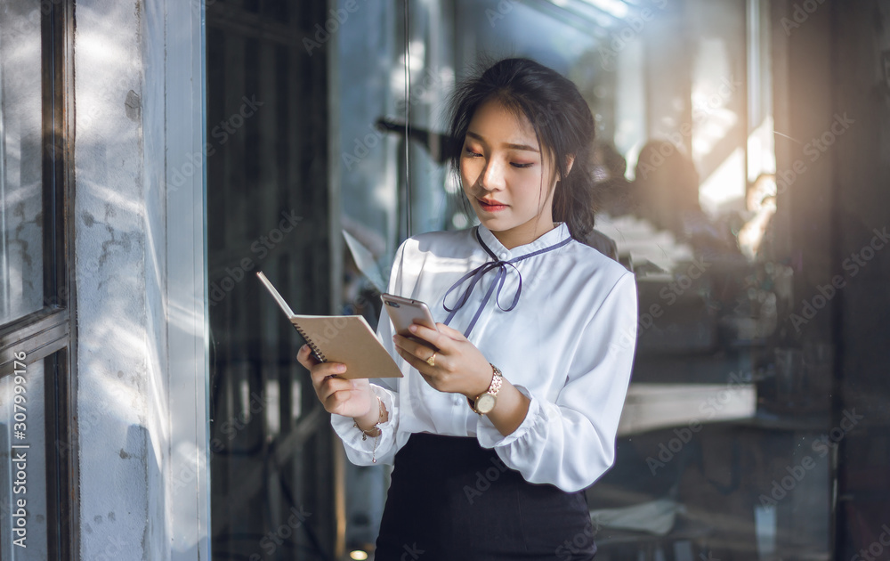 Young businesswoman checking information in note book and use internet from mobile phone to working Young businesswoman checking information in note book and use internet from mobile phone to working