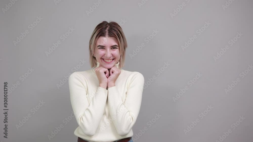 Pretty young caucasian lady keeping fists on chin with over gray background. Looks happy, funny wearing white sweater Pretty young caucasian lady keeping fists on chin with over gray background. Looks happy, funny wearing white sweater