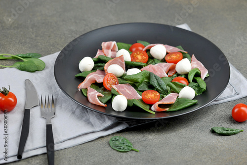 Salad with Parma ham, mozzarella cheese, cherry tomatoes and leafy spinach on a black plate.  In the frame a napkin and cutlery.  Close-up.