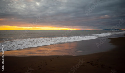 A sunset view of dark sand, waves, ocean and light reflected onto wet sand.