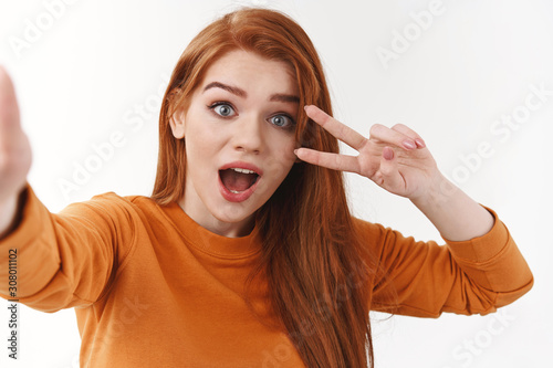 Surprised emotional cute redhead woman in orange sweater, holding camera with extended arm, taking selfie with funny photo filter, showing peace or victory sign, open mouth in awe or astonishment