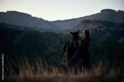 Beautiful young girl in a black dress hugs her black horse at sunset in the mountains