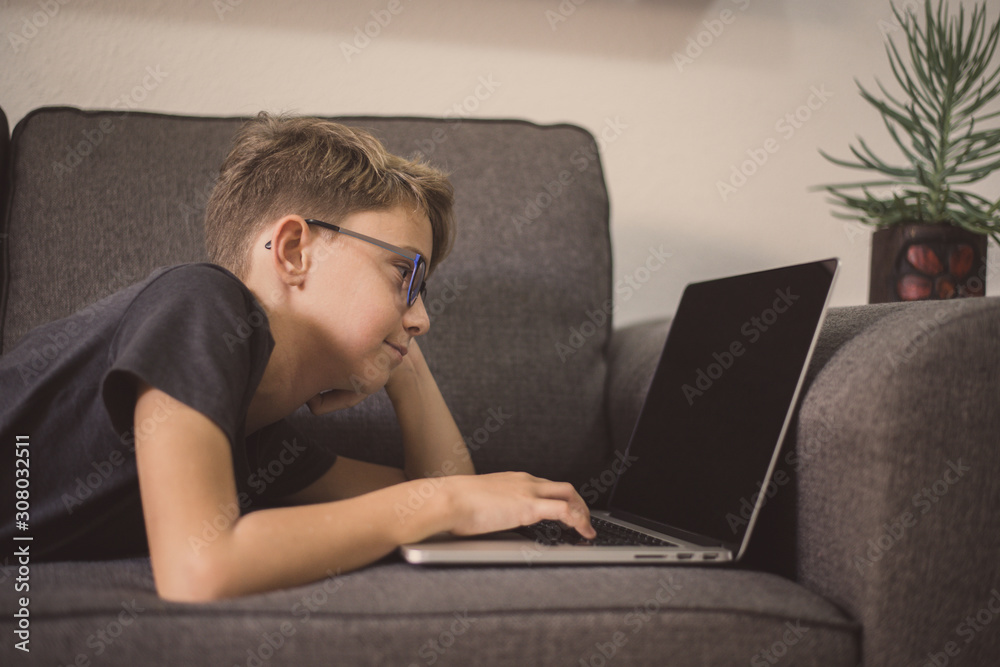 Young student doing homework lying on a sofa using laptop. Caucasian boy watching video online alone at home. Teen enjoying free time with computer. New technology education concept Soft focus on eyes Young student doing homework lying on a sofa using laptop. Caucasian boy watching video online alone at home. Teen enjoying free time with computer. New technology education concept Soft focus on eyes