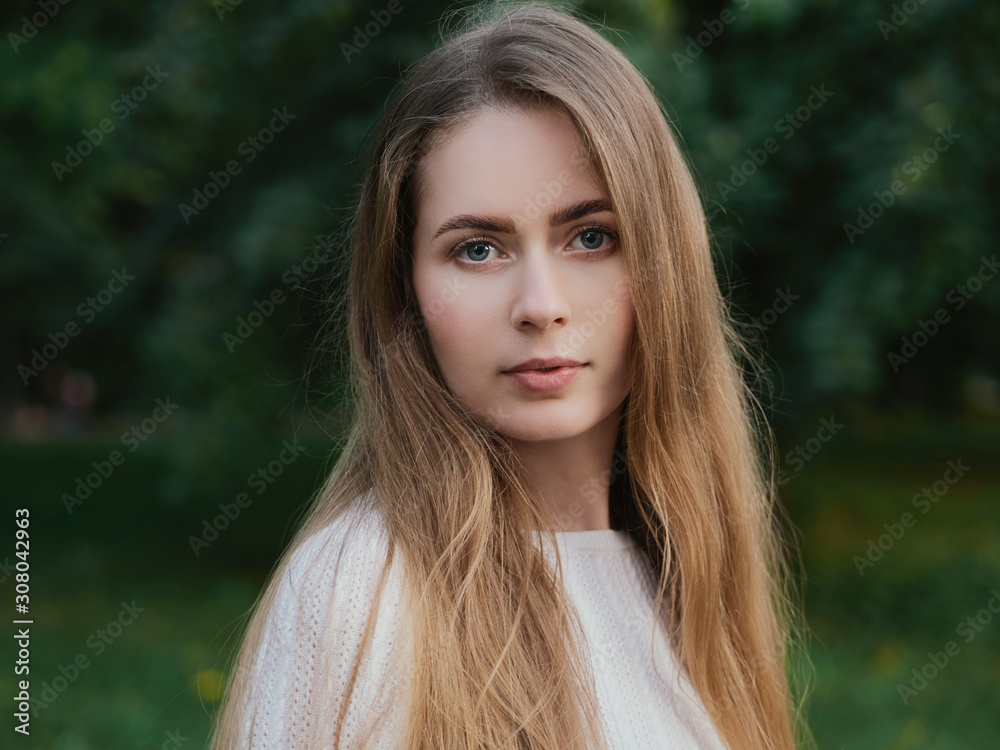 Close up headshot of adorable blonde haired charming smiling woman with blue eyes looking into camera in evening city park Close up headshot of adorable blonde haired charming smiling woman with blue eyes looking into camera in evening city park