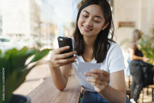 Carefree cheerful asian woman in urban cafe, sit near window at coffee table, hold smartphone, credit card, entder digits as send money friend, sharing bill with coworker, smiling as shopping online
