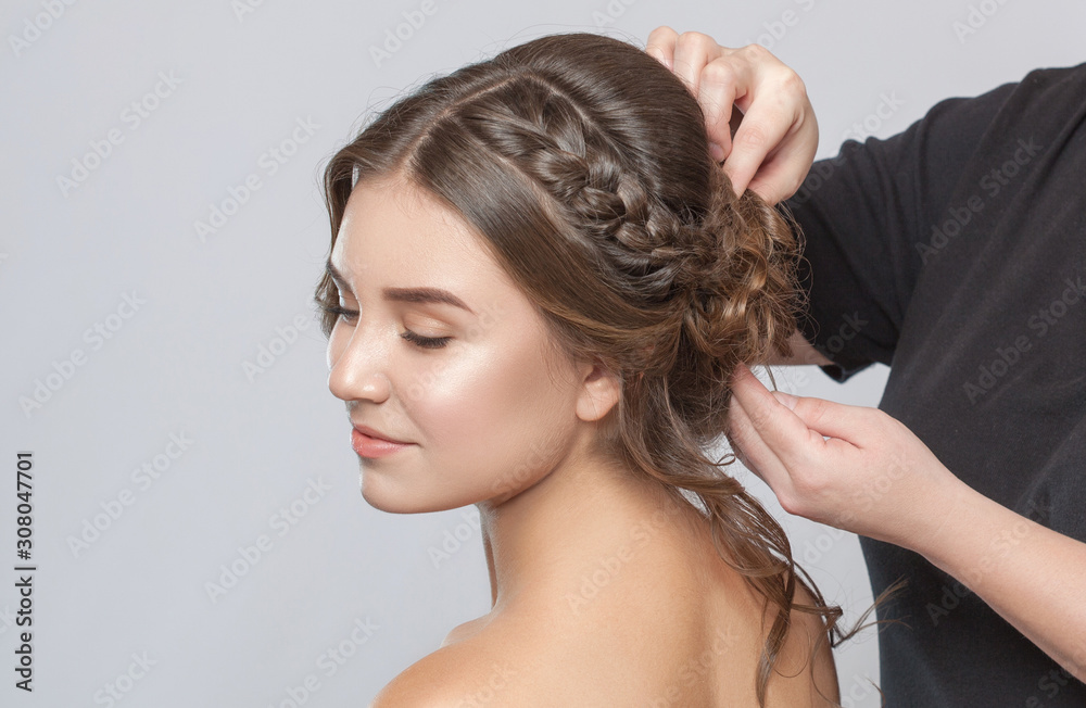 Naklejka premium Portrait of a beautiful sensual light brown haired woman with a wedding hairstyle in a beauty salon. The hairdresser does the hairstyle. Wedding hairstyle.