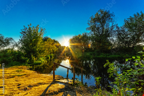 Beautiful sunset over the river. The sun shines through the trees. © VASILEVS
