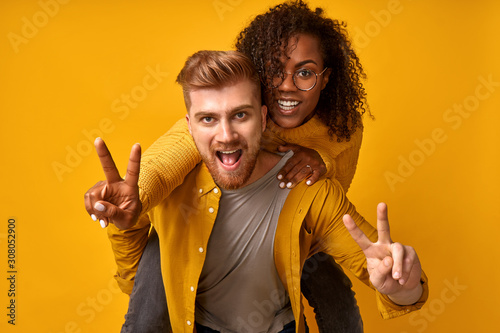 фотографія Photo of cheerful cute charming positive nice diverse couple showing you victory sign and giving a piggyback ride to joyful, wearing in monochrome orange, isolated over an orange color background