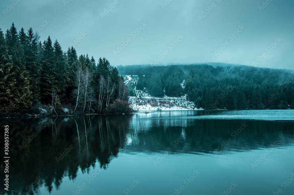 Winter mountain lake view with pine trees forest in the fog clouds and dramatic dark light. Walking in the nature mountains in winter times. Brocken, Harz National Park Mountains in Germany Winter mountain lake view with pine trees forest in the fog clouds and dramatic dark light. Walking in the nature mountains in winter times. Brocken, Harz National Park Mountains in Germany