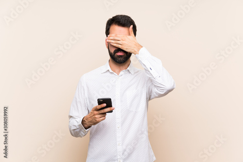 Young man with beard holding a mobile covering eyes by hands. Do not want to see something