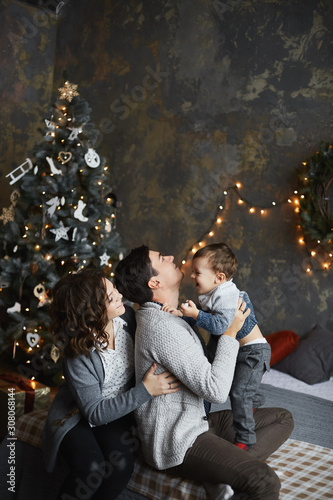Young parents and the baby boy hugs together in the interior decorated for New year. The father, mother and their little son have fun in the Christmas interior. Happy New Year and Merry Christmas © innarevyako